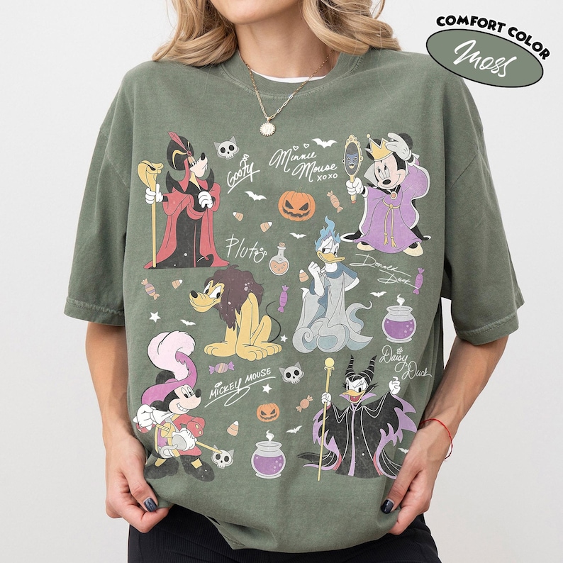 May include: A moss green short-sleeve t-shirt featuring a graphic print of various Disney characters in Halloween costumes, including Mickey Mouse, Minnie Mouse, Donald Duck, and Pluto, alongside pumpkins, candy, and text like Minnie Mouse XOXO.
