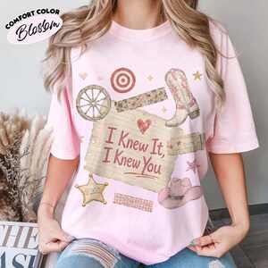 Op de afbeelding: Een lichtroze t-shirt met een grafische print van een gelinieerd briefje met de tekst I Knew It, I Knew You, omringd door illustraties van een wagenwiel, een cowboylaars, een sheriffster, een roze cowboyhoed, een doelwit en een ticket.