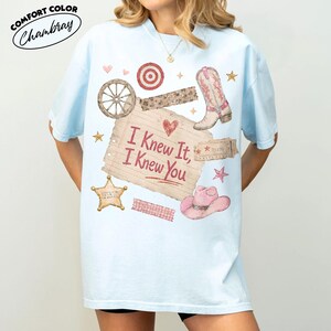 Op de afbeelding: Een lichtblauw t-shirt met een grafische print van een vel gelinieerd papier met de tekst "I Knew It, I Knew You" in het rood, omringd door illustraties van een cowboylaars, een roze hoed, een wagenwiel, een sheriffster en kleine hartjes.
