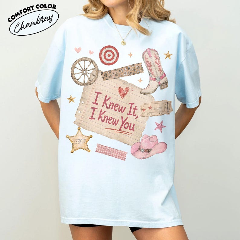 Op de afbeelding: Een lichtblauw t-shirt met een grafische print van een vel gelinieerd papier met de tekst "I Knew It, I Knew You" in het rood, omringd door illustraties van een cowboylaars, een roze hoed, een wagenwiel, een sheriffster en kleine hartjes.