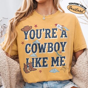 Peut inclure: Un t-shirt &agrave; manches courtes jaune moutarde avec le texte bleu fonc&eacute; "YOU'RE A COWBOY LIKE ME" et des motifs repr&eacute;sentant un chapeau de cowboy marron, une fus&eacute;e, une balle, des bottes, une &eacute;toile de sh&eacute;rif, des &eacute;toiles rouges et des nuages bleu clair.