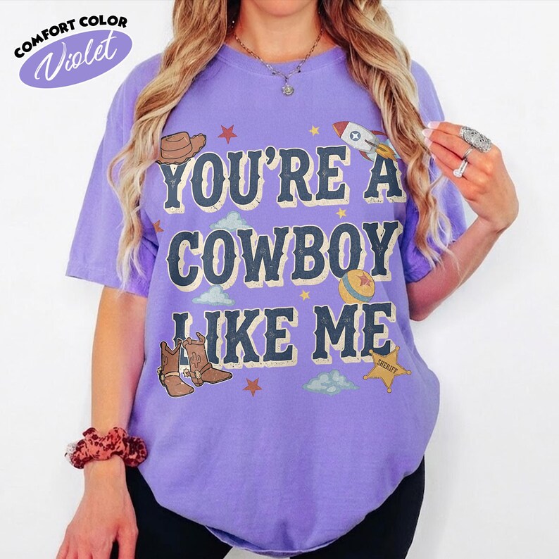 Peut inclure: Un t-shirt violet &agrave; manches courtes avec le texte "YOU'RE A COWBOY LIKE ME" dans une police vieillie. Le motif comprend des illustrations d'un chapeau de cowboy marron, d'une fus&eacute;e, d'une balle color&eacute;e, de bottes de cowboy et d'une &eacute;toile de sh&eacute;rif.