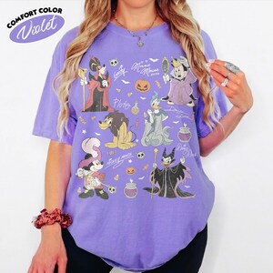 May include: A violet short-sleeve t-shirt featuring a graphic print of various cartoon characters dressed in Halloween costumes, including pumpkins, skulls, and potion bottles. The text Comfort Color Violet is printed in the upper left corner.