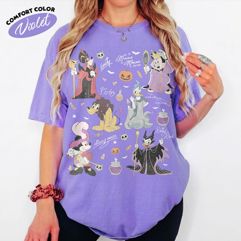 May include: A violet short-sleeve t-shirt featuring a graphic print of various cartoon characters dressed in Halloween costumes, including pumpkins, skulls, and potion bottles. The text Comfort Color Violet is printed in the upper left corner.