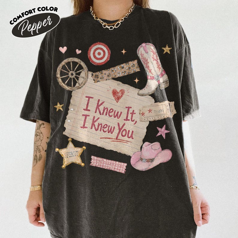 Op de afbeelding: Een zwart t-shirt met korte mouwen en een centrale afbeelding van een gelinieerd notitieblaadje met de tekst "I Knew It, I Knew You", omringd door illustraties van een cowboylaars, een roze cowboyhoed, een wagenwiel en kleine hartjes.