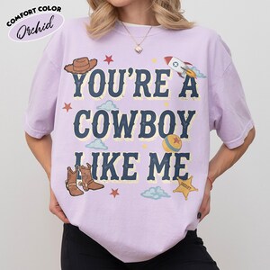 Peut inclure: Un t-shirt &agrave; manches courtes de couleur orchid&eacute;e avec le texte "YOU'RE A COWBOY LIKE ME" en lettres bleu fonc&eacute;. Le motif comprend un chapeau de cowboy marron, une fus&eacute;e, une balle color&eacute;e, des bottes de cowboy, une &eacute;toile de sh&eacute;rif et des &eacute;toiles rouges.