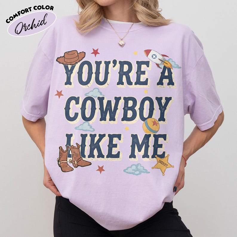 Peut inclure: Un t-shirt &agrave; manches courtes de couleur orchid&eacute;e avec le texte "YOU'RE A COWBOY LIKE ME" en lettres bleu fonc&eacute;. Le motif comprend un chapeau de cowboy marron, une fus&eacute;e, une balle color&eacute;e, des bottes de cowboy, une &eacute;toile de sh&eacute;rif et des &eacute;toiles rouges.