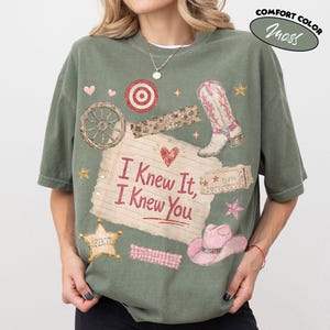 Op de afbeelding: Een mosgroen t-shirt met korte mouwen en een grafische print van een gelinieerd notitieblaadje met de tekst I Knew It, I Knew You. Het ontwerp bevat een cowboylaars, een roze cowboyhoed, een wagenwiel, een sheriffster en een doelwit.