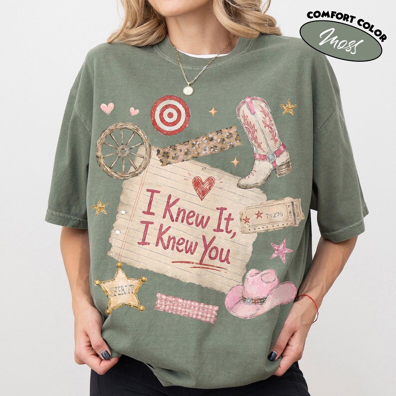 Op de afbeelding: Een mosgroen t-shirt met korte mouwen en een grafische print van een gelinieerd notitieblaadje met de tekst I Knew It, I Knew You. Het ontwerp bevat een cowboylaars, een roze cowboyhoed, een wagenwiel, een sheriffster en een doelwit.