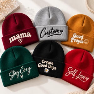 May include: Six knit beanies in maroon, gray, mustard yellow, dark green, black, and red. Each features embroidered text: mama with a heart, Custom with a heart, Good People with a smiley face, Stay Cozy with a heart, Create Good Days with a daisy, and Self Love with a heart.