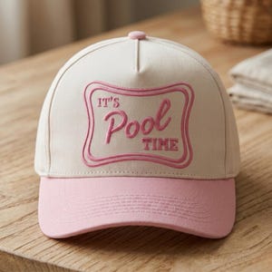 May include: A cream-colored baseball cap with a pink brim and a pink embroidered rectangular patch on the front. The patch features a double-line border and the text IT'S POOL TIME in a stylized pink font, centered on the front panel.