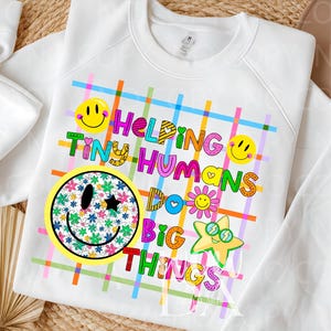 Puede incluir: Una sudadera blanca con un patrón de fondo de cuadros coloridos y el texto "HELPING TINY HUMANS DO BIG THINGS". El diseño incluye caras sonrientes amarillas, una cara sonriente con estampado floral, una estrella con gafas y flores.
