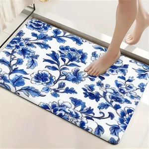 May include: A rectangular bath mat featuring a white background with a repeating blue floral pattern of blooming flowers and leafy vines. The mat has rounded corners and a smooth surface texture, displaying a consistent botanical design throughout.