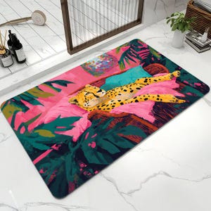 May include: A rectangular bath mat featuring a vibrant illustration of a cheetah resting on a pink cushion. The design includes deep green tropical foliage, a bright teal accent, and a colorful circular pattern against a bold pink background.