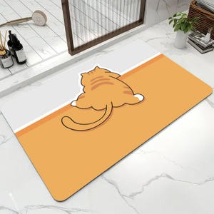 May include: A rectangular floor mat featuring a split design with a light gray upper section and a solid orange lower section. A cartoon illustration of an orange tabby cat is positioned in the center, shown from behind with its tail extended.