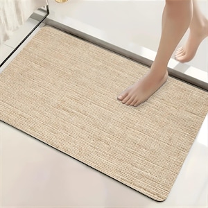 May include: A rectangular bath mat with a textured, woven beige surface that mimics the appearance of natural fibers. The mat features rounded corners and a flat, low-profile design, providing a stable surface for stepping out of a shower or bath.