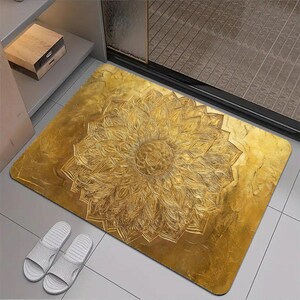 May include: A rectangular floor mat featuring a metallic gold finish and a large, intricate embossed mandala pattern centered on the surface. The design consists of numerous layered, petal-like shapes radiating outward from a circular core.