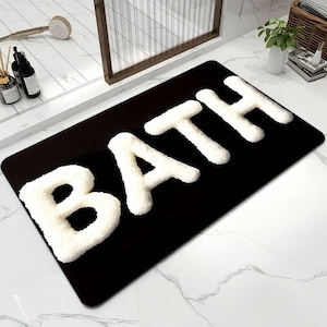 May include: A rectangular bath mat featuring a solid black background with the word BATH spelled out in large, raised, white, textured, fuzzy letters. The mat has rounded corners and a soft, plush appearance, contrasting against the dark base.
