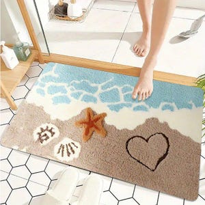 May include: A rectangular bath mat featuring a beach-themed design with light blue ocean waves, white sea foam, and tan sand. The mat includes a textured orange starfish, two white seashells, and a heart shape outlined in the sand.