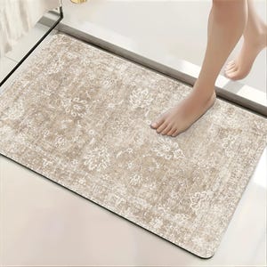May include: A rectangular bath mat featuring a distressed, faded beige and cream color palette with an intricate, symmetrical floral pattern reminiscent of traditional rug designs. The mat has a soft, textured surface and rounded corners.