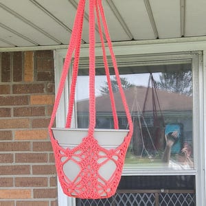 Pode incluir: Um suporte de plantas em crochê na cor coral neon brilhante com um design de nós intrincado e quatro cordões de suspensão longos que se unem em um laço superior, segurando um vaso de flores de plástico cinza claro.