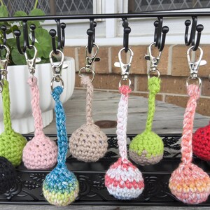 May include: A collection of nine crocheted ball keychains hanging from a black metal rack. Each keychain features a round, textured yarn sphere attached to a braided cord and a silver-toned metal lobster clasp. The colours include black, lime green, light pink, blue, beige, variegated pink and white, bright green, variegated orange and pink, and solid red.