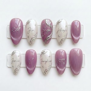 May include: A set of ten artificial nails in various designs. Some nails are solid lavender, while others are white or lavender with silver accents and text. The text includes the names "RM", "Jin", "Suga", "j-hope", "Jimin", "V", "Jungkook", and "BTS".