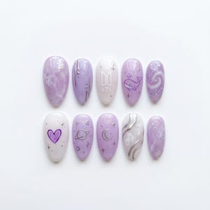 May include: A set of ten almond-shaped press-on nails in shades of lavender, purple, and white. The designs feature silver metallic accents, a purple heart, a whale illustration, a planet, a crescent moon, and the BTS logo in white text.