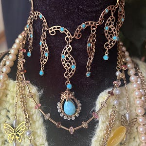 May include: A layered necklace set featuring a gold-tone choker with filigree accents and small turquoise stones, a central teardrop pendant with a blue stone and white beads, and additional chains adorned with pearls, pink crystals, and a gold butterfly.