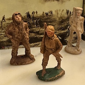 May include: Three vintage-style military figurines standing on a surface. One figure wears a flight suit and goggles, another carries a rifle, and the third stands in a uniform. The figures have a textured, weathered brown and tan finish.