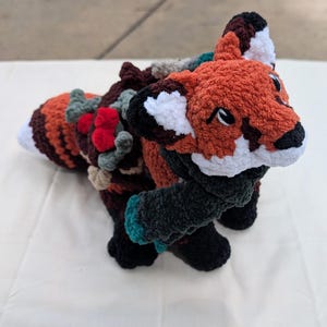 May include: A handmade, plush fox toy with orange, white, black, and brown yarn. The fox has a detailed face, white ear tips, and a striped tail. The body features decorative yarn embellishments in various colors. The toy is designed to stand upright.