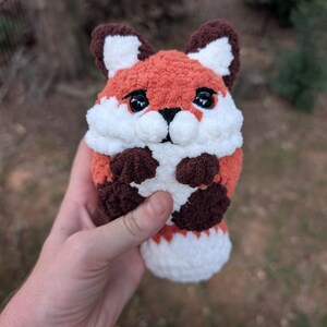 May include: A small, round, handmade crochet fox plush toy featuring a textured orange body, white chest and face accents, dark brown ears, and dark brown paws. The fox has large, glossy black safety eyes and a small black stitched nose.