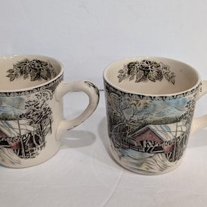 May include: Two cream-colored ceramic mugs featuring a detailed transferware scene of a red covered bridge in a snowy landscape. The interior rim of each mug is decorated with a black and white botanical pattern of leaves and grapes.