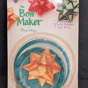 May include: The Bow Maker by Bow Magic is a teal plastic tool for creating ribbon bows, packaged on a white card featuring images of red, green, and orange bows with text reading Decorations, Floral Designs, and Gift Wrap.