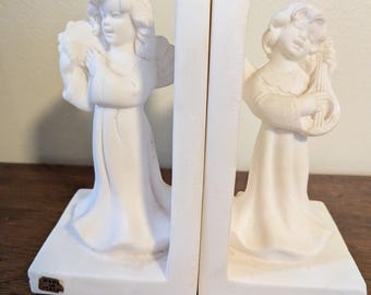 May include: A pair of white matte bookends featuring sculpted angels in long robes. One angel holds a tambourine, while the other plays a stringed instrument. A small rectangular sticker on the base reads Made in Italy in black text.