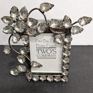 May include: A rectangular metal photo frame featuring a dark finish and clear faceted crystal accents arranged in a floral and vine pattern. The center insert displays the text Two By Two Photo Frame TWO'S COMPANY in black lettering on a white background.