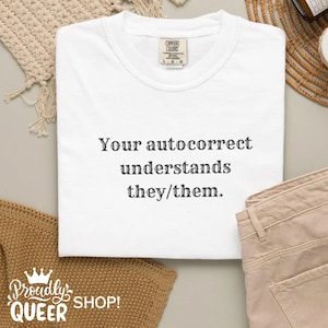 May include: White Comfort Colors t-shirt with the text "Your autocorrect understands they/them." The shirt has a crew neck and a small tag on the upper chest. The shirt is folded and next to beige trousers.