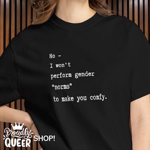 Può includere: T-shirt nero con testo bianco: "No - I won't perform gender 'norms' to make you comfy." La maglietta presenta anche il testo "Proudly QUEER SHOP!" in un carattere stilizzato.