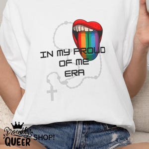 May include: A white t-shirt featuring a graphic of an open mouth with a rainbow-coloured tongue. The text "IN MY PROUD OF ME ERA" is printed in black letters. A faint, light grey outline of a rosary with a cross is visible behind the text.