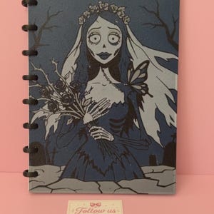 Puede incluir: Un cuaderno rectangular con cubierta azul oscuro que presenta una ilustración gráfica de una novia esquelética con velo y corona de flores sosteniendo un ramo de rosas. El cuaderno tiene encuadernación de discos negros. Una tarjeta dice Follow us on INSTAGRAM.