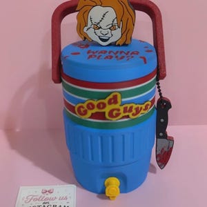 Puede incluir: Una nevera portátil de plástico azul con asa roja y grifo amarillo. Presenta una banda horizontal a rayas rojas, blancas y verdes con el texto Good Guys, una figura de Chucky en la parte superior y un pequeño dije de cuchillo en el asa.