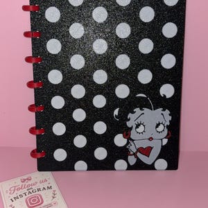 Puede incluir: Un cuaderno negro con lunares blancos y textura brillante, que presenta un personaje de dibujos animados con cabello negro y pendientes rojos sosteniendo un corazón rojo. El cuaderno está encuadernado con discos de plástico rojo.