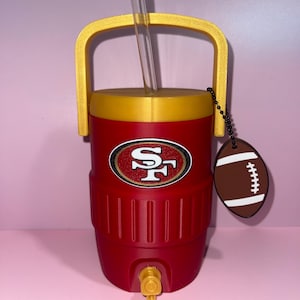 May include: A red and gold insulated beverage cooler with a handle, a clear straw, and a spigot at the base. The front features a San Francisco 49ers logo, and a small brown football-shaped charm hangs from the handle on a black beaded chain.