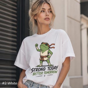 Op de afbeelding: Een wit t-shirt met korte mouwen en een afbeelding van een groene kikker in een vechtsportuniform met een rode hoofdband en riem. Onder de kikker staat de tekst "STRONG TODAY BETTER TOMORROW" in zwarte en groene letters.