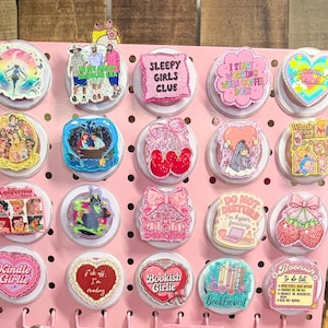 May include: A pink display board showcasing various round pop sockets. Designs include text like "Sleepy Girls Club", "Bookish Girlie", and illustrations of characters, bows, and strawberries. The pop sockets are on a pink display board.