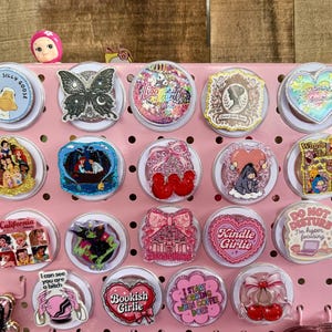 Puede incluir: Un tablero perforado rosa que muestra varios soportes circulares para teléfono con diseños diversos, incluyendo personajes de dibujos animados, texto como 'Kindle Girlie' y 'Bookish Girlie', mariposas brillantes, cerezas y patrones coloridos en cuadrícula.