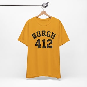 Peut inclure: Un t-shirt à manches courtes jaune moutarde avec le texte "BURGH" dans une police noire arquée au-dessus du nombre "412" dans une police noire assortie. Le lettrage présente un aspect vieilli et usé sur le devant du vêtement.
