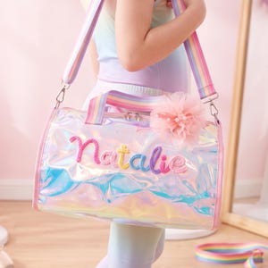 May include: A cylindrical duffel bag made of iridescent, holographic material featuring the name Natalie embroidered in colorful yarn letters. The bag includes a rainbow-striped shoulder strap, matching top handles, and a soft pink tulle pom-pom.