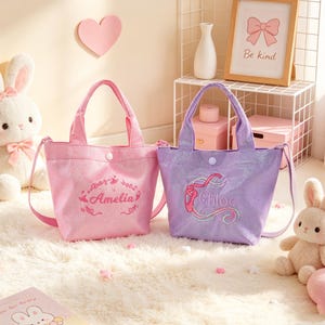 May include: Two glittery tote bags with shoulder straps and snap closures. The pink bag features the name Amelia in cursive script with a ballerina graphic. The purple bag features the name Chloe in cursive script with a ballet slipper graphic.