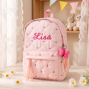May include: A light pink quilted backpack featuring a diamond pattern with small embroidered bows. The name Lisa is embroidered in dark pink script on the front. A pink bow charm and tassel are attached to the side zipper pull.
